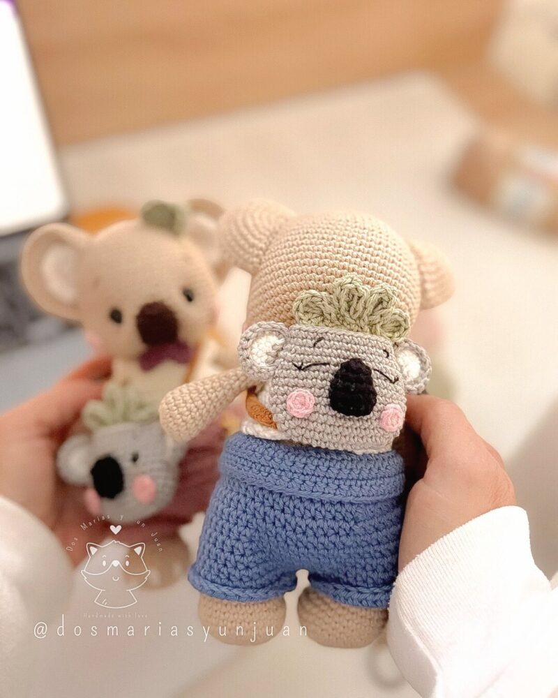 Rear view of Benny the crochet koala showing the mini koala backpack on his back. Blurred Benna figure in the background adds depth.
