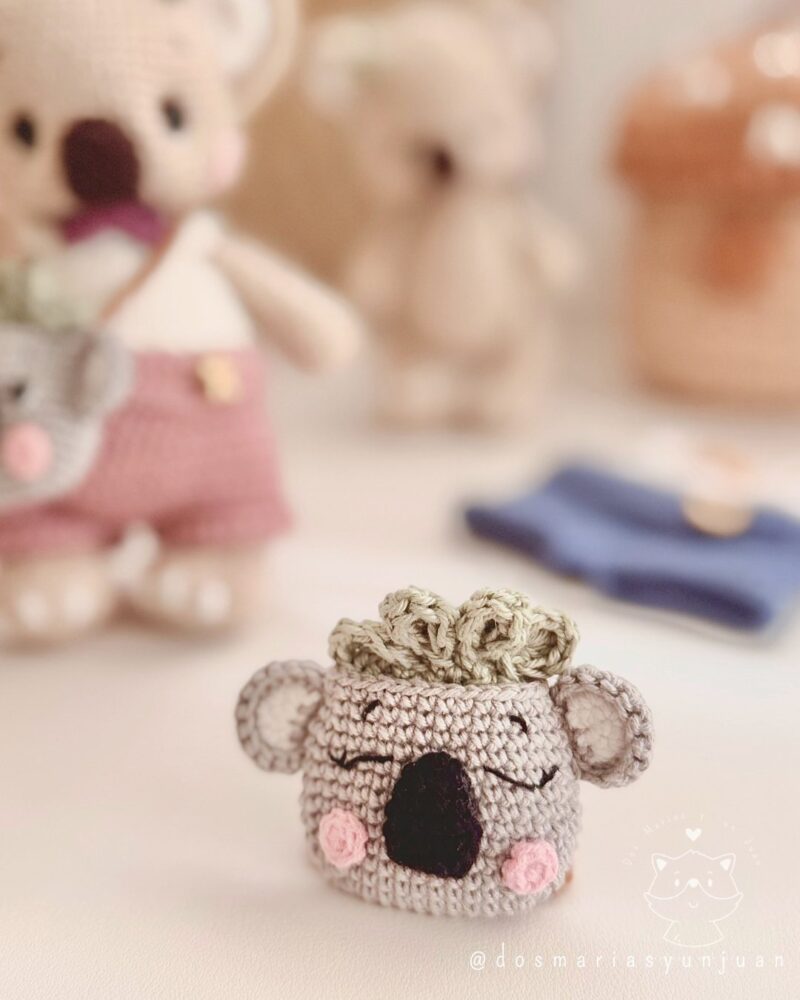Close-up of the crochet koala backpack with embroidered sleepy eyes, pink cheeks and leaf detail. Blurred Benna and Benny in the background.