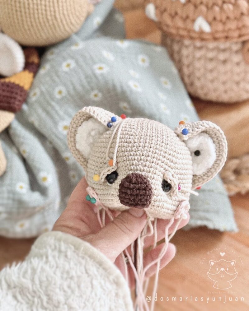 Close-up of a crochet koala head in progress, with colorful pins and loose yarn ends. A behind-the-scenes view of the handmade process by Dos Marías y un Juan.