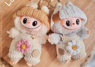 Two handmade Labubu dolls wearing crochet overalls with flower appliqués and beanies. Beige wooden background.