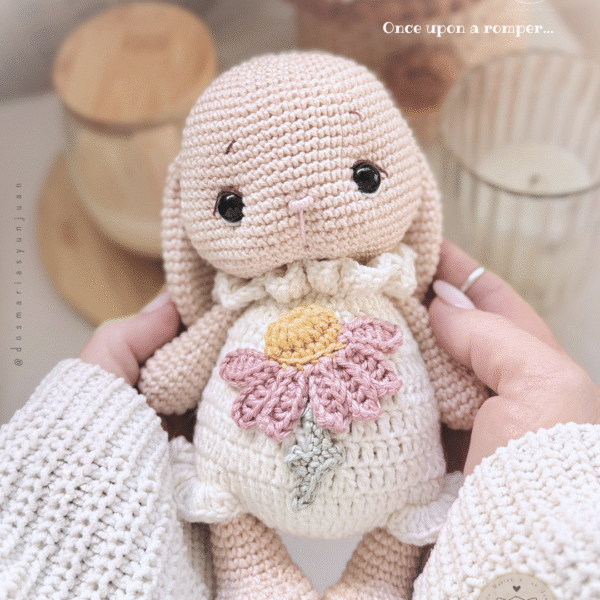 Front view of Miss Flopps Bun amigurumi, wearing a ruffled romper and pink daisy applique; hands holding the bunny close up.