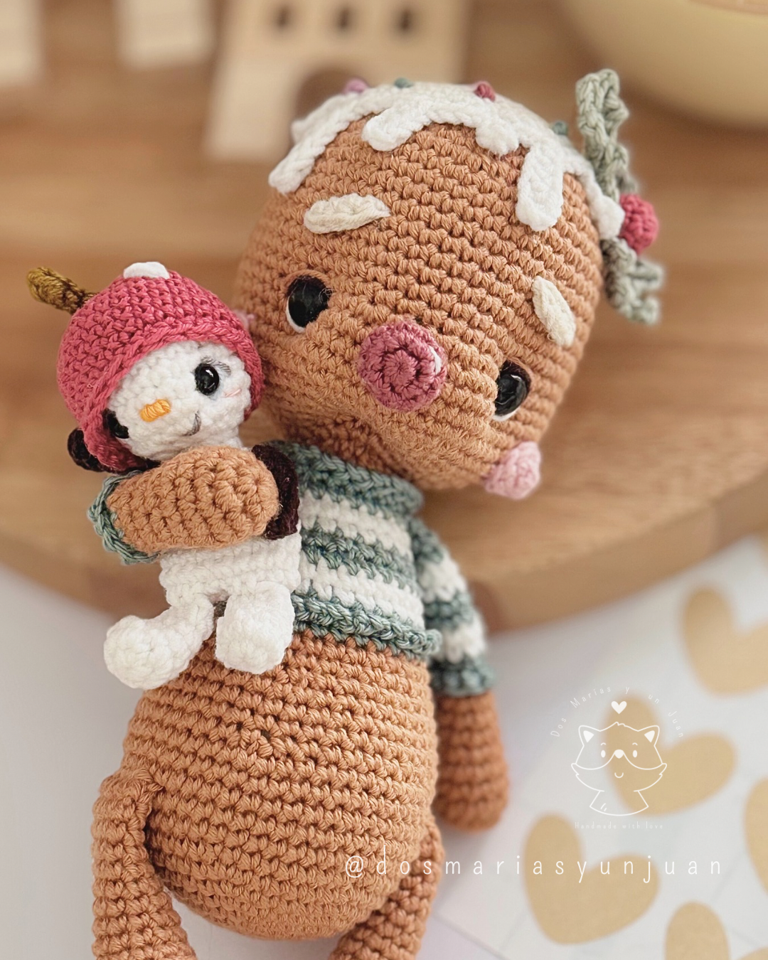 Close-up of Candito gingerbread amigurumi hugging Snowy Micro Snuggler, with Christmas decor behind.