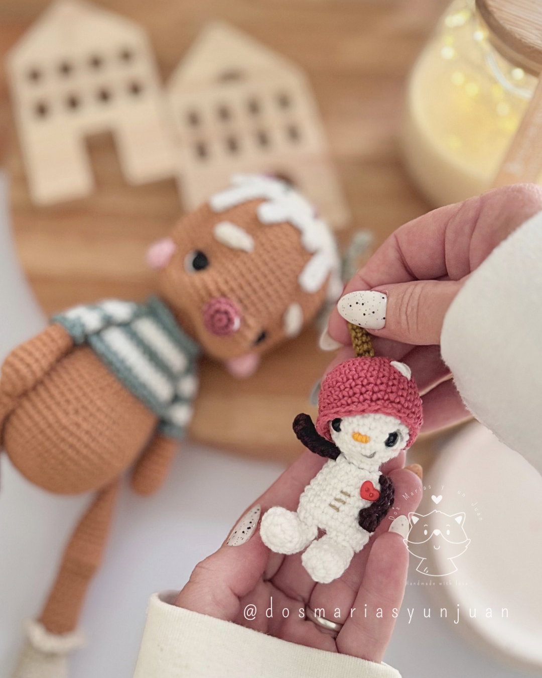 Close-up of Snowy Micro Snuggler amigurumi held by hand, with blurred Candito gingerbread doll in the background.